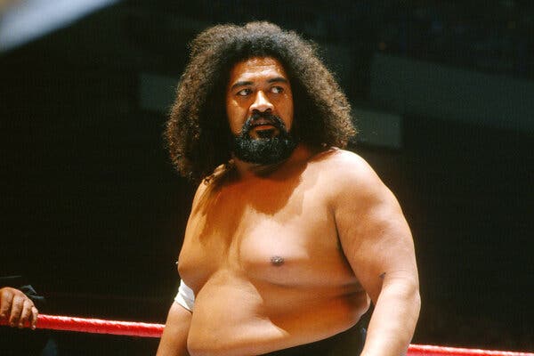 Sika Anoa’i looks on during a wrestling match. He has dark curly hair and a beard.