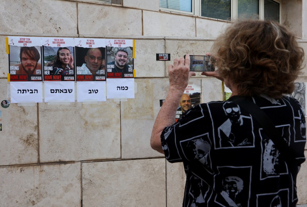 A woman using her mobile phone to photograph 'Home Now' posters showing the portraits of four rescued Israeli hostages on a wall in Tel Aviv amid ongoing conflict.