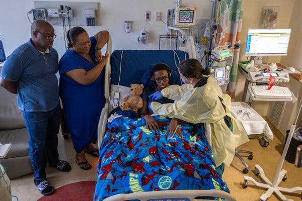 A view looking down on Kendric’s hospital bed, which is decorated with Spider-Man, as a nurse attends to him and his parents stand at his side.