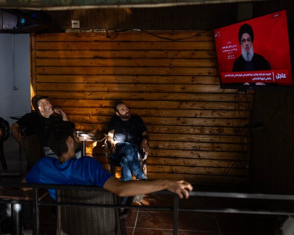 Several men sit in a darkened room, watching a man speak on television. 