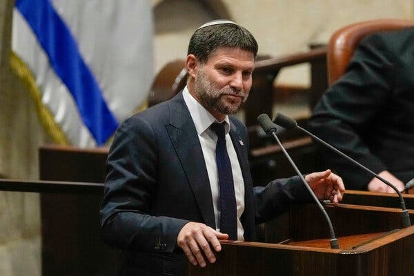 Bezalel Smotrich standing at a pedestal with two microphones. He is wearing a dark gray suit and a dark tie.