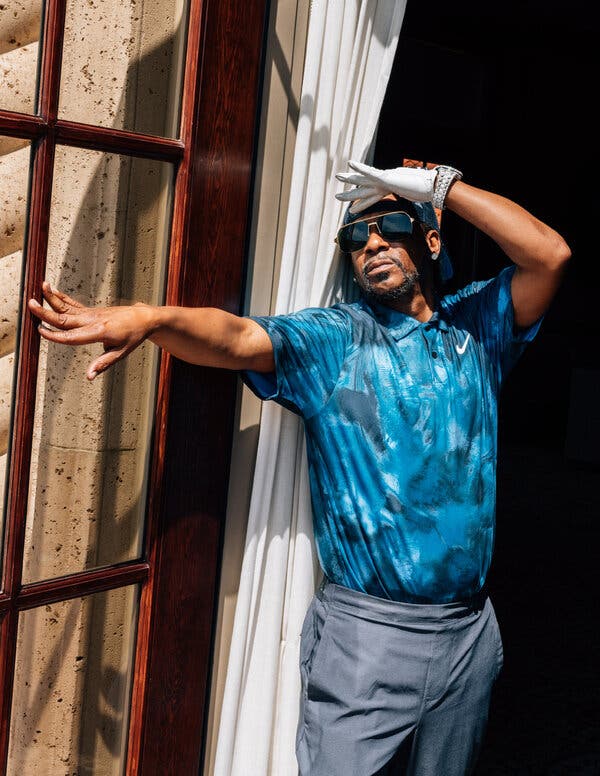 Standing in a doorway next to a curtain, Katt Williams, in golf clothes and sunglasses, extends one arm while putting a gloved hand up to his forehead as if shading his eyes from the sun.